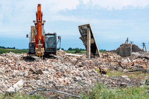 Orange crusher excavator stands on ruins of old brick farm building and crushes concrete structures.