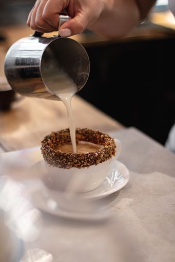 A high angle shot of a barista making coffee with milk in a white cup partially covered in chocolate and nut crumbs