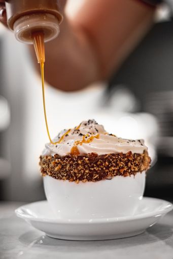 A high angle shot of a barista making coffee with caramel sauce and whipped cream in a white cup covered in chocolate spread and nut crumbs