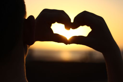 A silhouette shot of hands making a heart shape over a background of a sunset