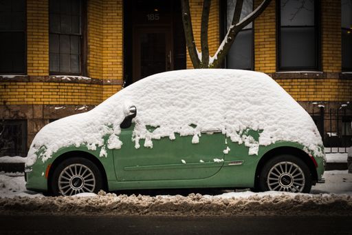 A green car half-covered with snow parked in front of a building with yellow walls in the background