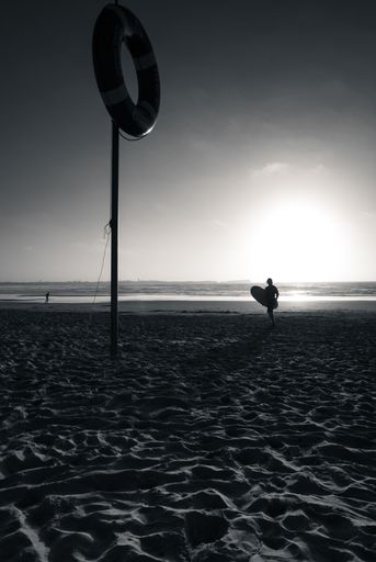 A vertical grayscale shot of a surfer's silhouette near a lifebuoy at a beach