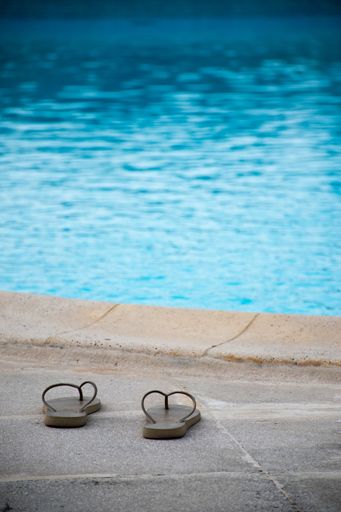A vertical shot of dark male slippers on a stone-ground in front of pure blue water of a pool