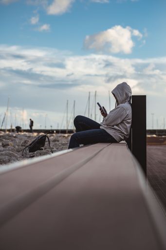 A vertical shot of a woman wearing an oversized hoodie looking at her phone sitting on a bench