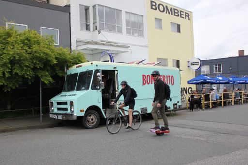 A young man and woman cycling and skateboarding near a food truck in Vancouver, Canada.