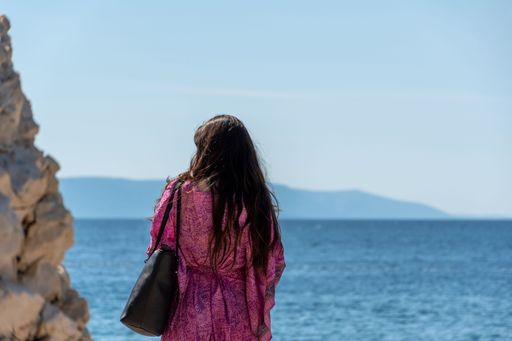 A selective focus shot of a girl in a dress with a bag standing against the sea