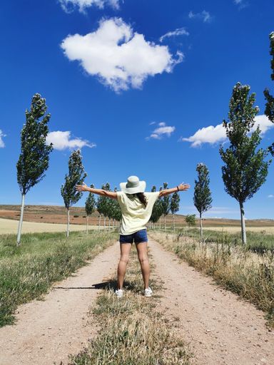 A vertical shot of a female from behind standing on the rural road with trees on both sides on a sunny day