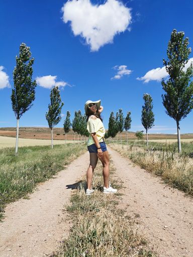 A vertical shot of a female standing on the rural road with trees on both sides on a sunny day