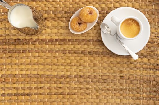 A high angle shot of a cup of coffee, donuts, and a pitcher of milk on a woven surface