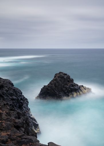 A vertical long exposure shot of water flow around large rock formations in a sea