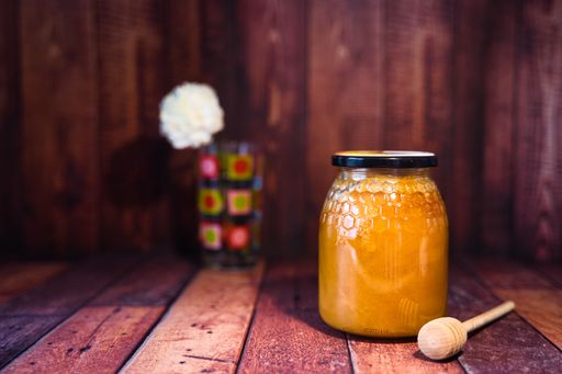 A jar of bee honey with a honey dipper on a wooden surface with a blurred flower in a glass in the background