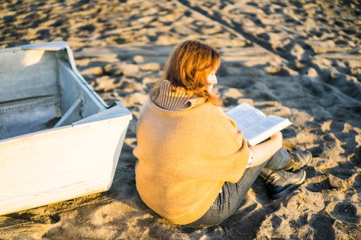 A back view of a ginger caucasian female sitting and reading a book on the sand at a beach