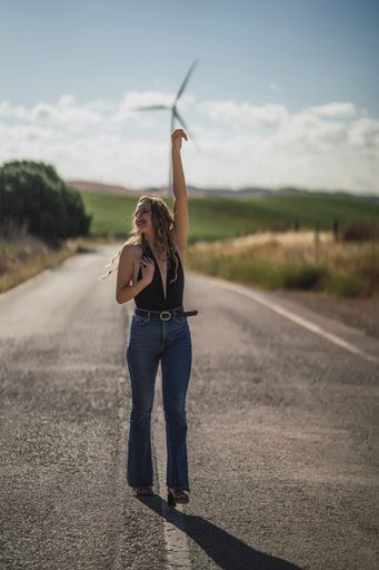 A vertical shot of a young Caucasian female walking on a road near a field with wind turbines