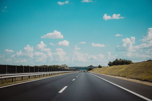 An empty asphalted road with white markings, railings and trees beside it under a blue sky with clouds