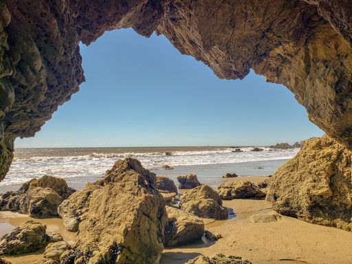A scenic view of ocean waves crushing against a sandy beach seen from a rocky cave