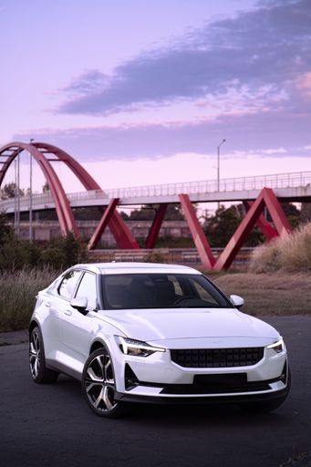A vertical shot of a white Polestar 2 in front of a red bridge at sunset