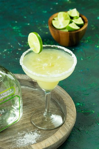 A vertical of a glass of Margarita cocktail on a wooden plate next to a tequila bottle and sliced lime