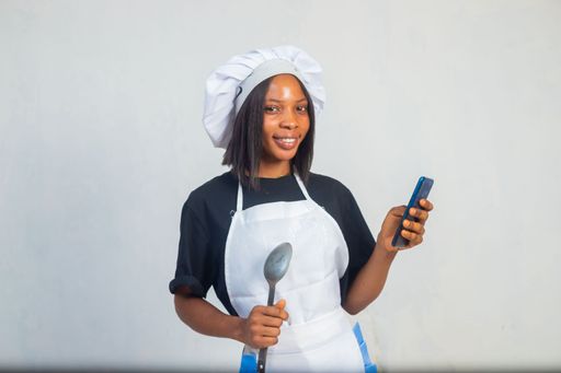 An African-American female in a white apron and hat holding a big spoon and a smartphone against a white background