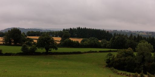 Panoramic shot of farmland landscape with green field, dense forest on hills in Aveyron, France under cloudy sky