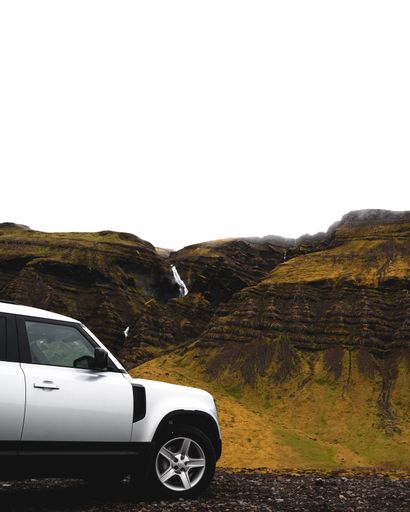 A vertical shot of a white car parking in front of mountains with waterfalls, Iceland