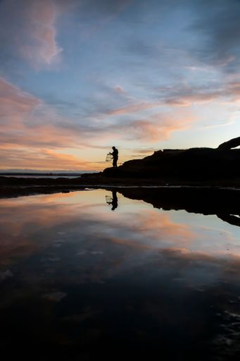 A silhouette shot of a person standing on a rocky beach while fishing in New South Wales, Australia