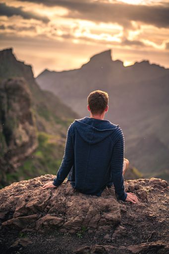 A Young Caucasian male sitting on rocks with a landscape of green mountains and sunset sky