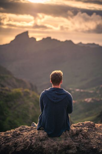 A Young Caucasian male sitting on rocks with a landscape of green mountains and sunset sky