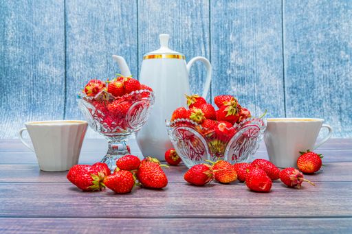 Red ripe strawberries lie in two crystal vases and on a wooden table. A coffee pot and two cups stand side by side.