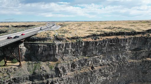 A bird's eye view of a bridge going over a steep cliff with an open field in the background