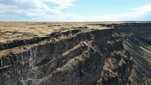 A bird's eye view of a steep cliff covered with an open field with the sky in the horizon