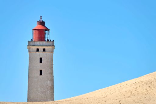 An old lighthouse with weathered walls and people on it surrounded by sand dunes at the beach under a pale blue sky with no clouds