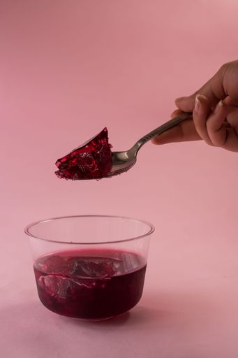 A vertical shot of a hand with a spoon picking up purple jelly from a bowl on a pink background