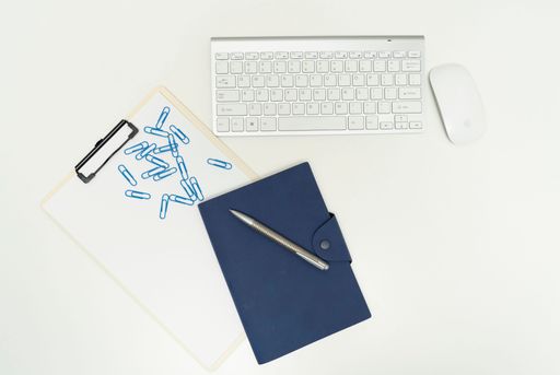 A top view of a mouse and keyboard next to a pen on top of a clipboard with blue paperclips