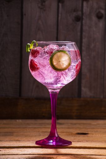 A vertical shot of a glass of pink tonic and gin with raspberries and lime on a wooden table