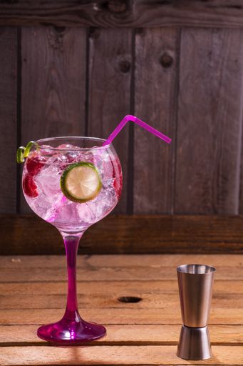 A vertical shot of a glass of pink tonic and gin with raspberries and lime on a wooden table