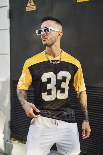 A vertical shot of a young European tattooed hip male in a yellow and black sporty outfit, sunglasses, and metal chain posing in front of a garage