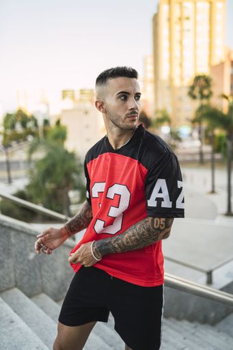 A vertical shot of a young European hip male in a sporty outfit with red and black shorts and shirt, tattoos, and metal chain standing on the stairs