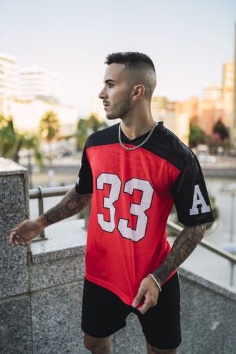 A vertical shot of a young European hip male in a sporty outfit with red and black shorts and shirt, tattoos, and metal chain standing on the stairs
