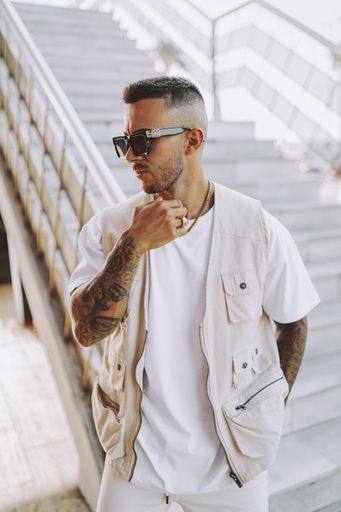 A vertical shot of a young tattooed male in a vest and sunglasses standing near the staircase