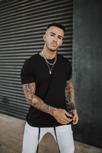 A vertical shot of a young European tattooed hip male in a black shirt and white ripped jeans, and metal chain standing in front of a garage