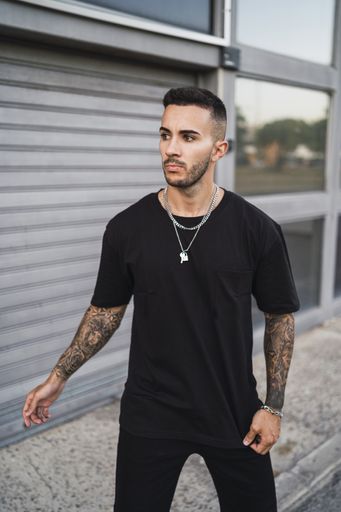 A vertical shot of a young European tattooed hip male in a black sporty outfit and a metal chain standing in front of a garage