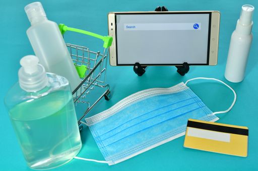 A miniature shopping trolley, a medical mask, alcogel sprays, a card and a smartphone on a blue surface