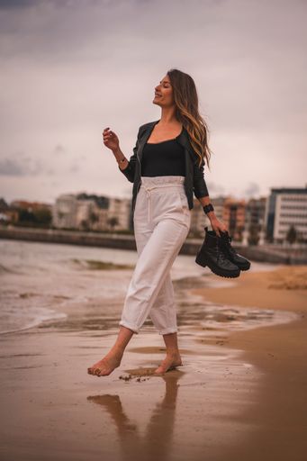 A vertical shot of a young stylish female walking barefoot on a beach at sunset holding her shores
