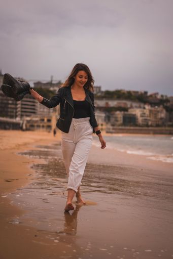 A vertical shot of a young stylish female walking barefoot on a beach at sunset holding her shores