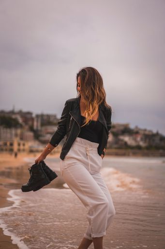 A vertical shot of a young stylish female walking barefoot on a beach at sunset holding her shores