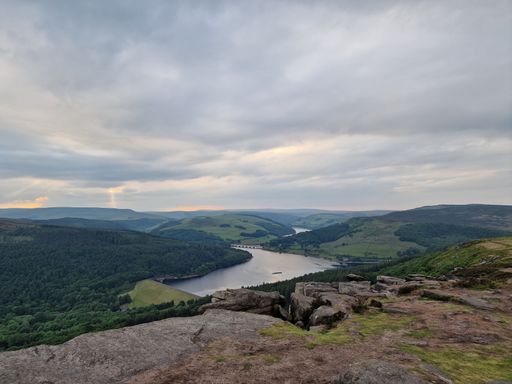 A scenic shot of a green landscape and a narrow lake from a rocky mountain with an overcast sky