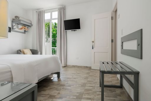 White bedroom with lateral view of a bed, place for suitcases, tv and balcony door with view of a tree