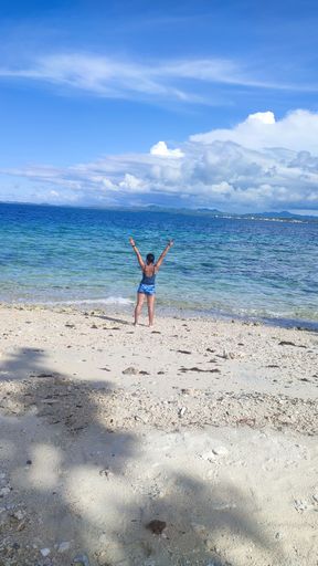 A vertical shot of a woman standing at the seashore with the ocean and blue sky in the background