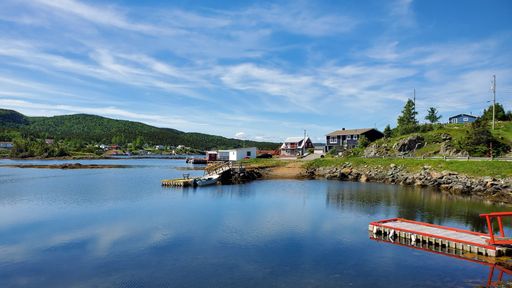 A sea coast, with houses and green trees, with red wooden pontoon on the shallow water, and a forested mountain in the background