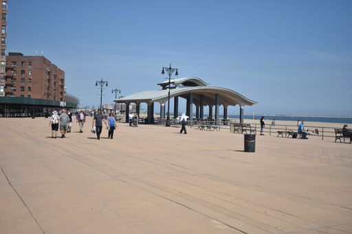 Brighton Beach near Coney Island, Brooklyn boardwalk, with buildings and seating structure in background, people walking, blue sky